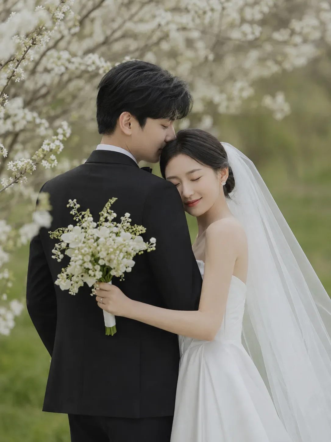 Hana resting her head on Josh's shoulder among white blossoms, holding a white bouquet