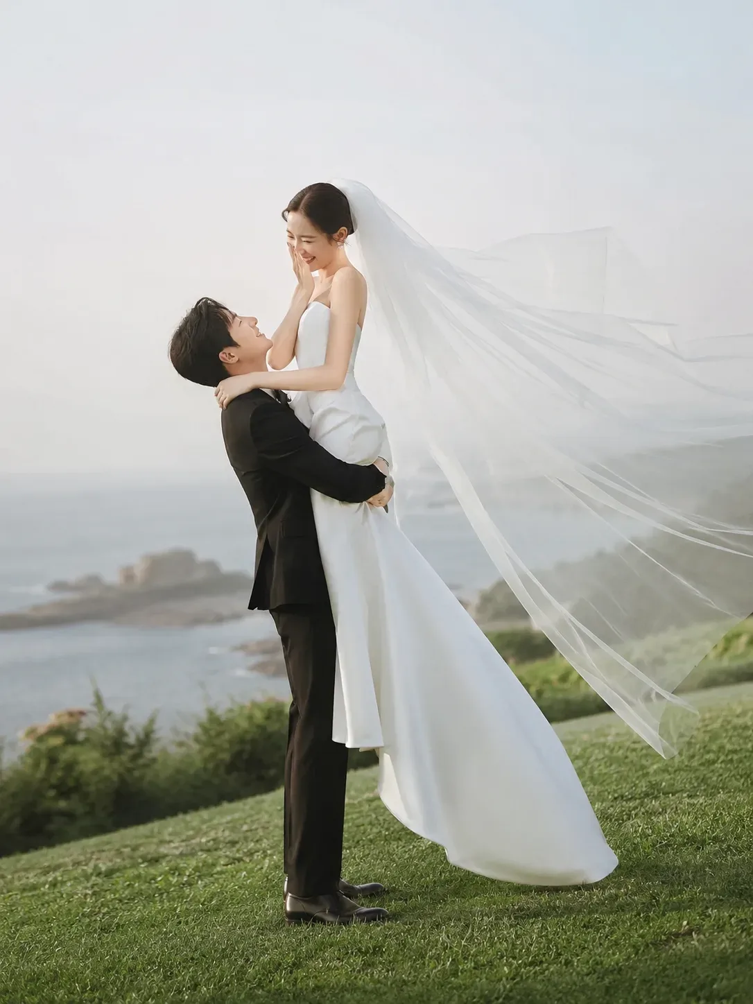 Josh lifting Hana high on a hillside above the sea, her veil flying